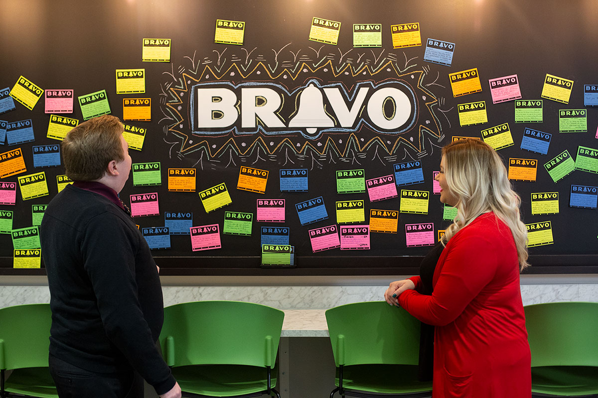 man and woman standing in front of a chalkboard that says BRAVO, surrounding by post it notes